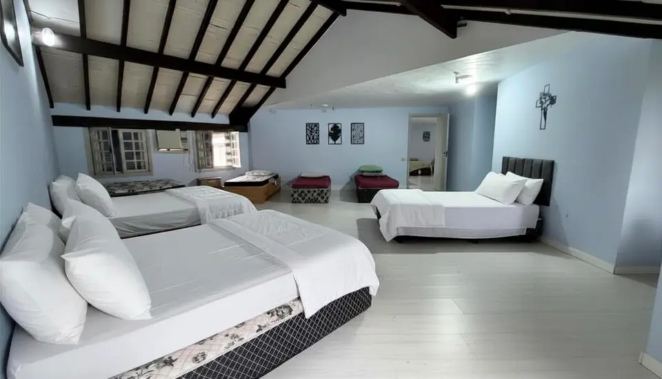 Quarto 5 da mansão, com camas de casal e de solteiro sob telhado com estrutura de madeira aparente